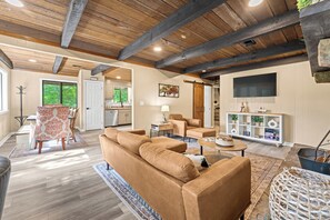 A spacious living area with a large brown sofa, a dining table with chairs, a kitchen in the background, and a television mounted on the wall. - Cozy 2BR for 4 w/ BBQ Grill, Mins to Attractions! (Gatlinburg)