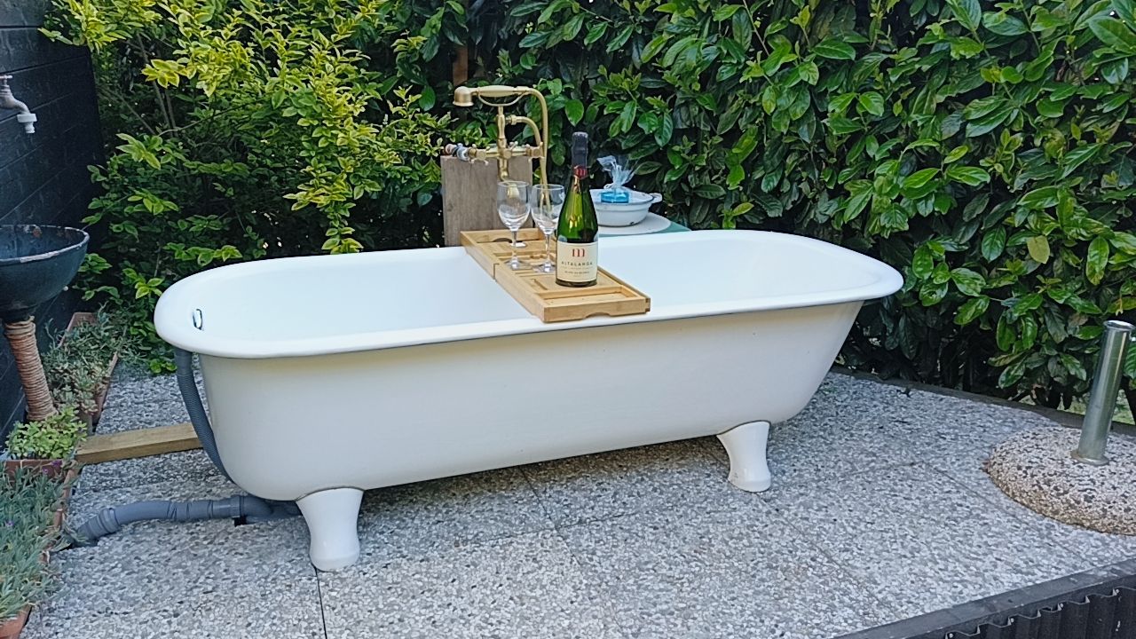 Una vasca da bagno indipendente con una bottiglia di vino e bicchieri su un vassoio, posata su una superficie in pietra con vista su un giardino.