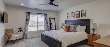A bedroom with a large bed, a ceiling fan, a window with curtains, a bedside table with a lamp, and a small chair.