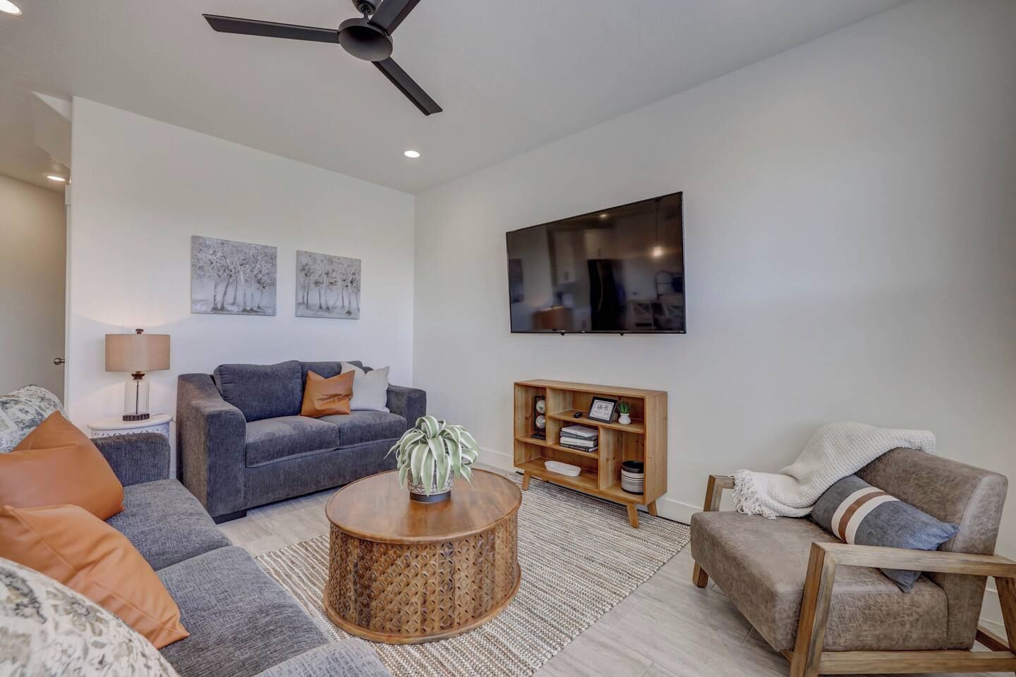 A modern living room with a grey sofa, a round coffee table, a wooden side table, a television mounted on the wall, and a ceiling fan.