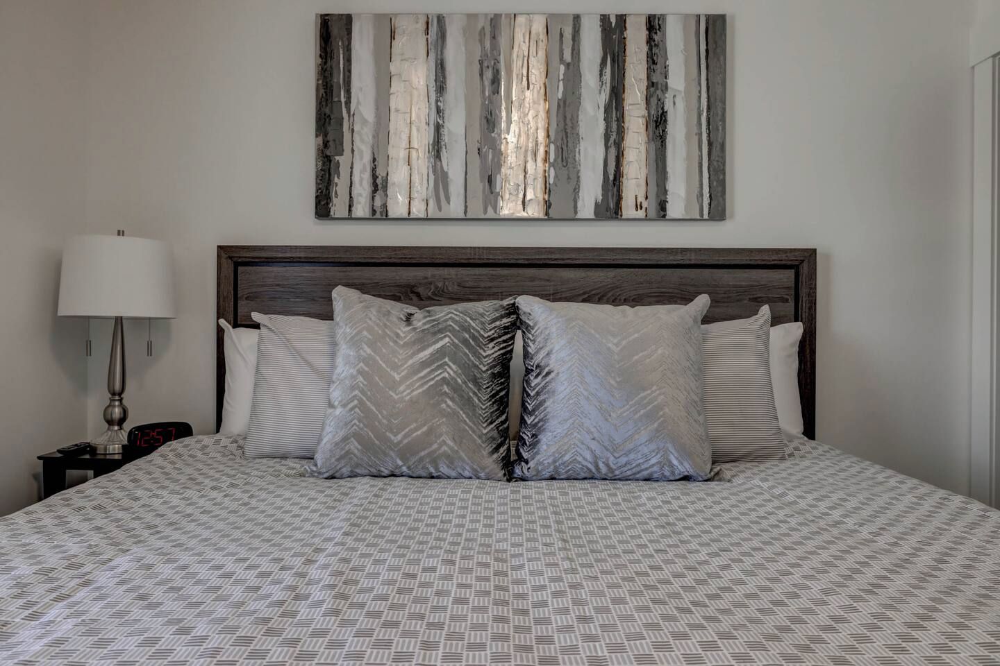 A neatly made bed with patterned bedding, a wooden headboard, and a wall-mounted abstract painting.