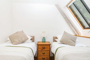 Ein Schlafzimmer mit zwei Betten, einem Nachttisch, einer Lampe und einem Fenster mit Blick auf Grünflächen.