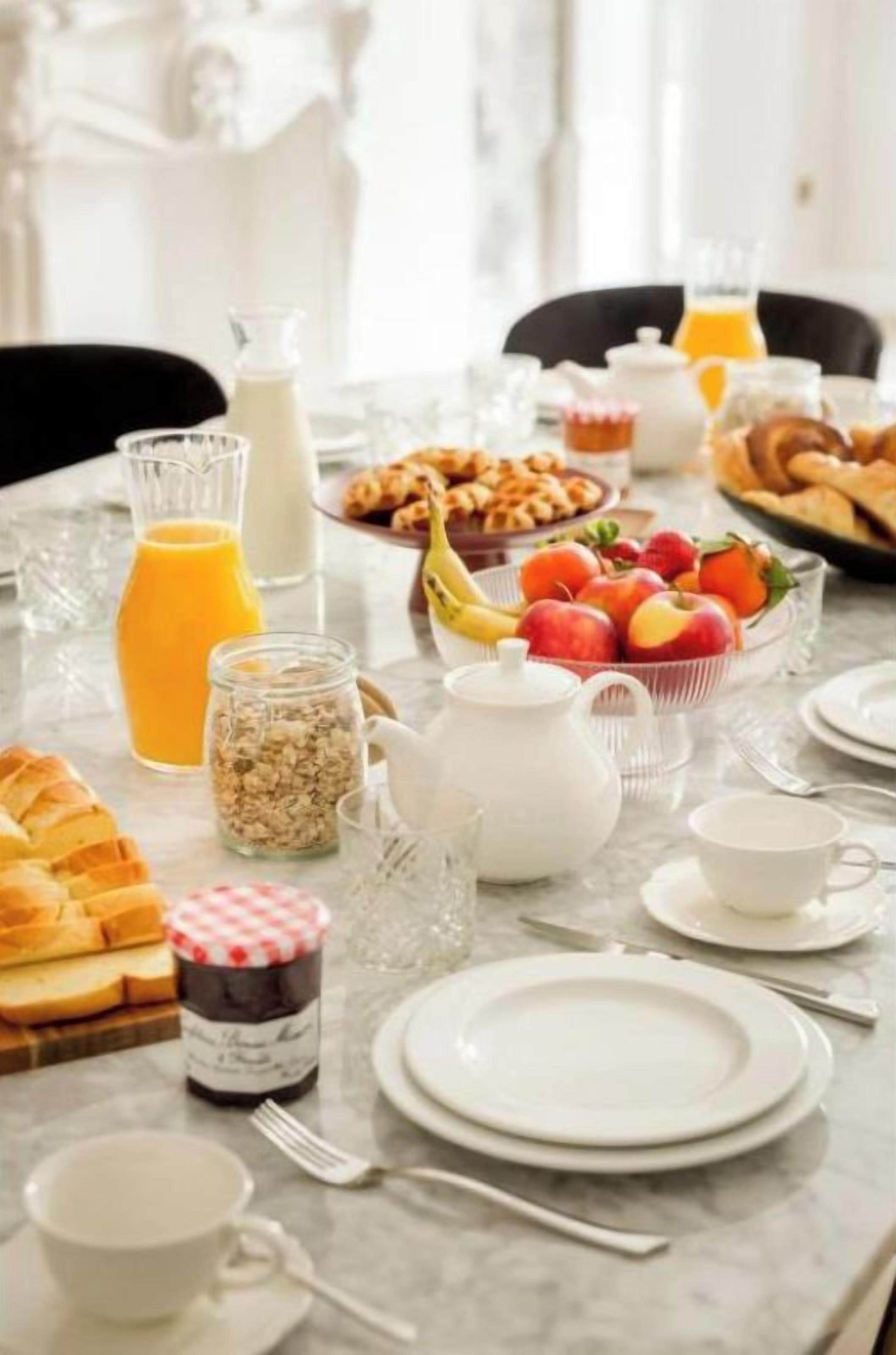 A breakfast table set with plates, cups, a jug of orange juice, a bowl of fruit, and a bottle of milk.