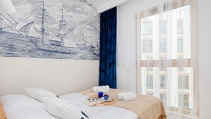 Ein ordentlich bezogenes Bett mit Kissen und einem Tablett mit Tasse und Gläsern, ein großes Fenster mit durchsichtigen Vorhängen und ein Wandbild mit Segelschiffen.