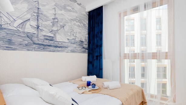 A neatly made bed with pillows and a tray with a cup and glasses, a large window with sheer curtains, and a wall mural of sailing ships. - FourInnaGB doNotBook (Guildford)