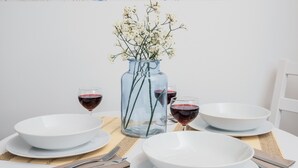 A dining table with white plates, bowls, and glasses of red wine, a clear glass vase with white flowers, and a woven placemat. - FourInnaGB doNotBook (Guildford)