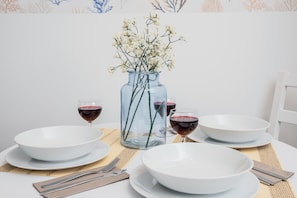 A dining table with white plates, bowls, and glasses of red wine, a clear glass vase with white flowers, and a woven placemat. - FourInnaGB doNotBook (Guildford)