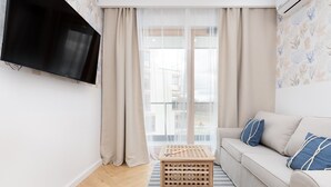 Ein Wohnzimmer mit einer Couch, einem an der Wand montierten Fernseher, einem Holztisch und einem Fenster mit durchsichtigen Vorhängen.