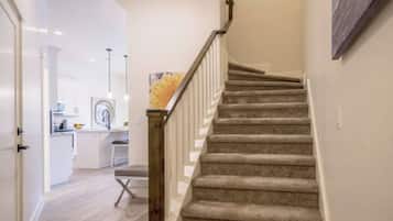 Eine Treppe mit Holzgeländer, das zu einer Zwischenebene führt, ein Küchenbereich mit weißen Schränken sowie ein Esstisch mit Stühlen.