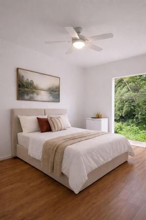 Et soveværelse med seng, ventilator, et natbord og et vindue med udsigt til grøn vegetation.