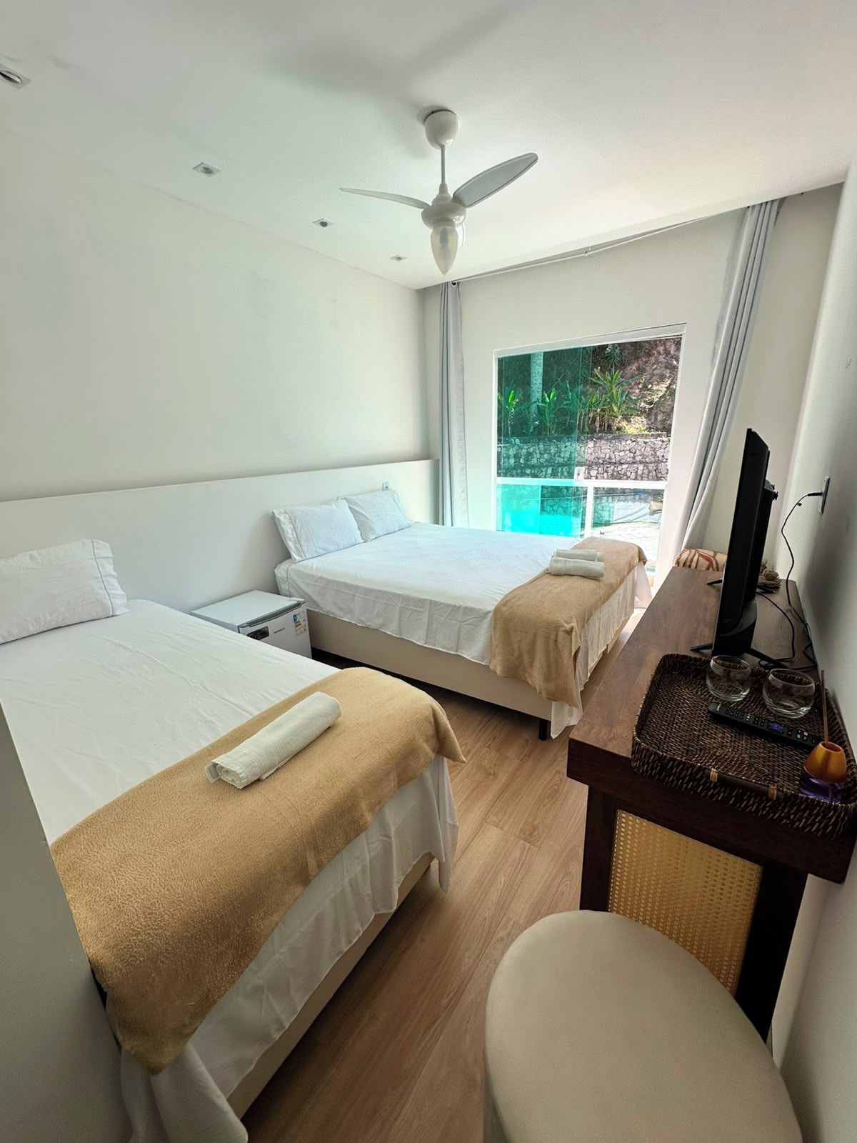 A hotel room with two beds, a desk with a TV, and a window with a view of greenery.