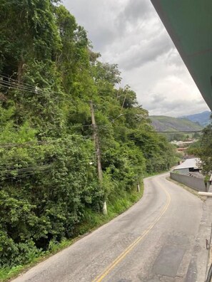 A curving road with dense greenery on one side and a clear view of a hilly landscape in the distance.