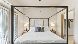 Una cama con dosel y ropa de cama blanca, dos mesitas de noche con lámparas, un cuadro enmarcado en la pared y una puerta a la derecha.