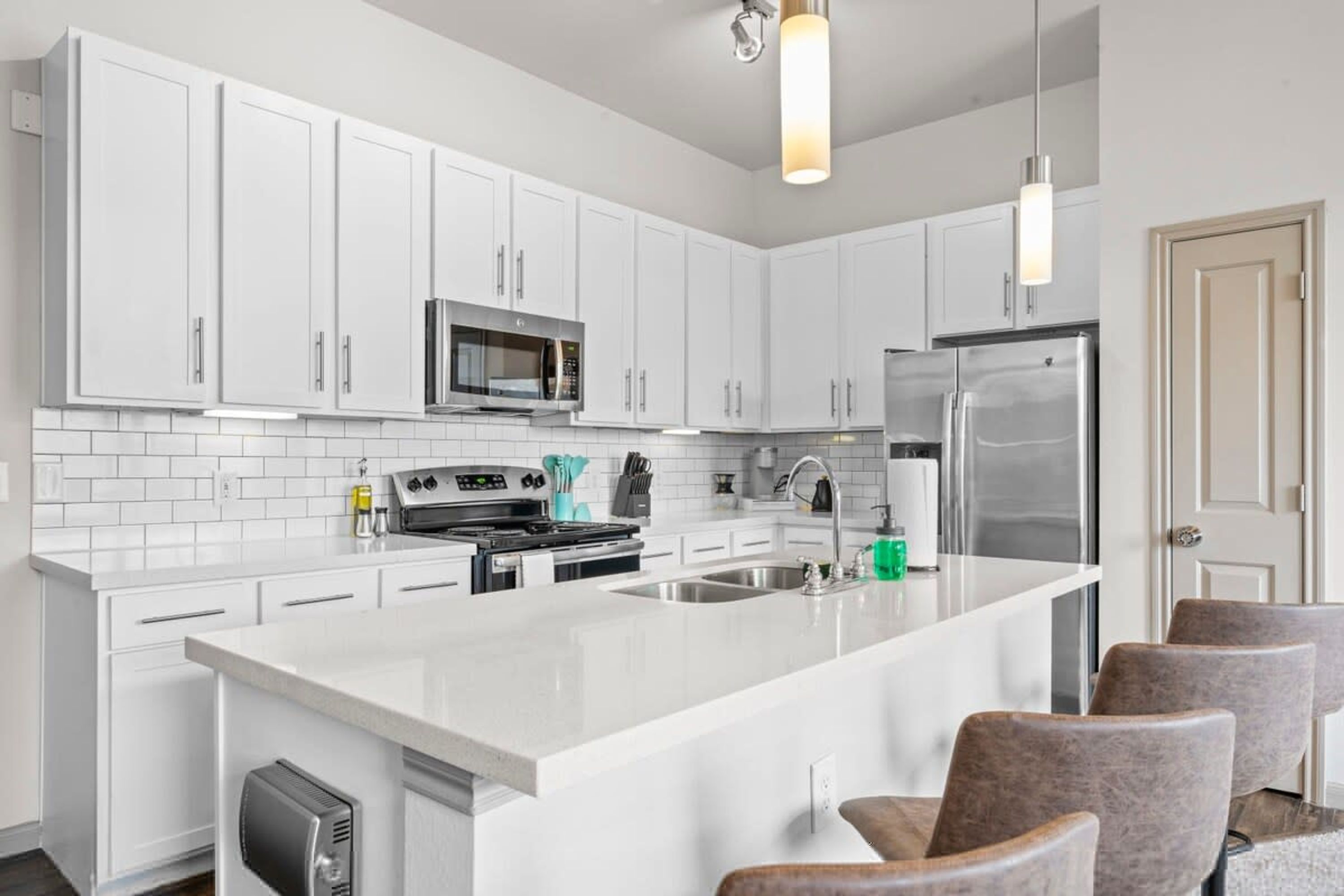 Una cocina moderna con muebles blancos, electrodomésticos de acero inoxidable, una isla central con asientos e iluminación colgante.