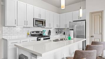 Una cocina moderna con muebles blancos, electrodomésticos de acero inoxidable, una isla central con asientos e iluminación colgante.