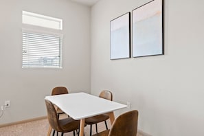 Una habitación con una mesa blanca y cuatro sillas, una ventana con persianas, y dos cuadros en la pared.