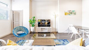 Uma sala de estar moderna com uma TV de tela plana, uma área de jantar com mesa de vidro e um espaço de estar com uma poltrona azul e um sofá.