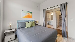 A bedroom with a bed, nightstands, a painting, and a bathroom visible through an open door. - Elegant 2BR King Suite Fast Wifi 8 min to DT (Austin)