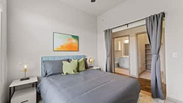 Un dormitorio con cama, mesitas de noche, un cuadro y un baño visible a través de una puerta abierta.