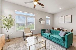 A living room with a teal sofa, a glass coffee table, a ceiling fan, and a view of the outdoors through large windows. - Elegant 2BR King Suite Fast Wifi 8 min to DT (Austin)