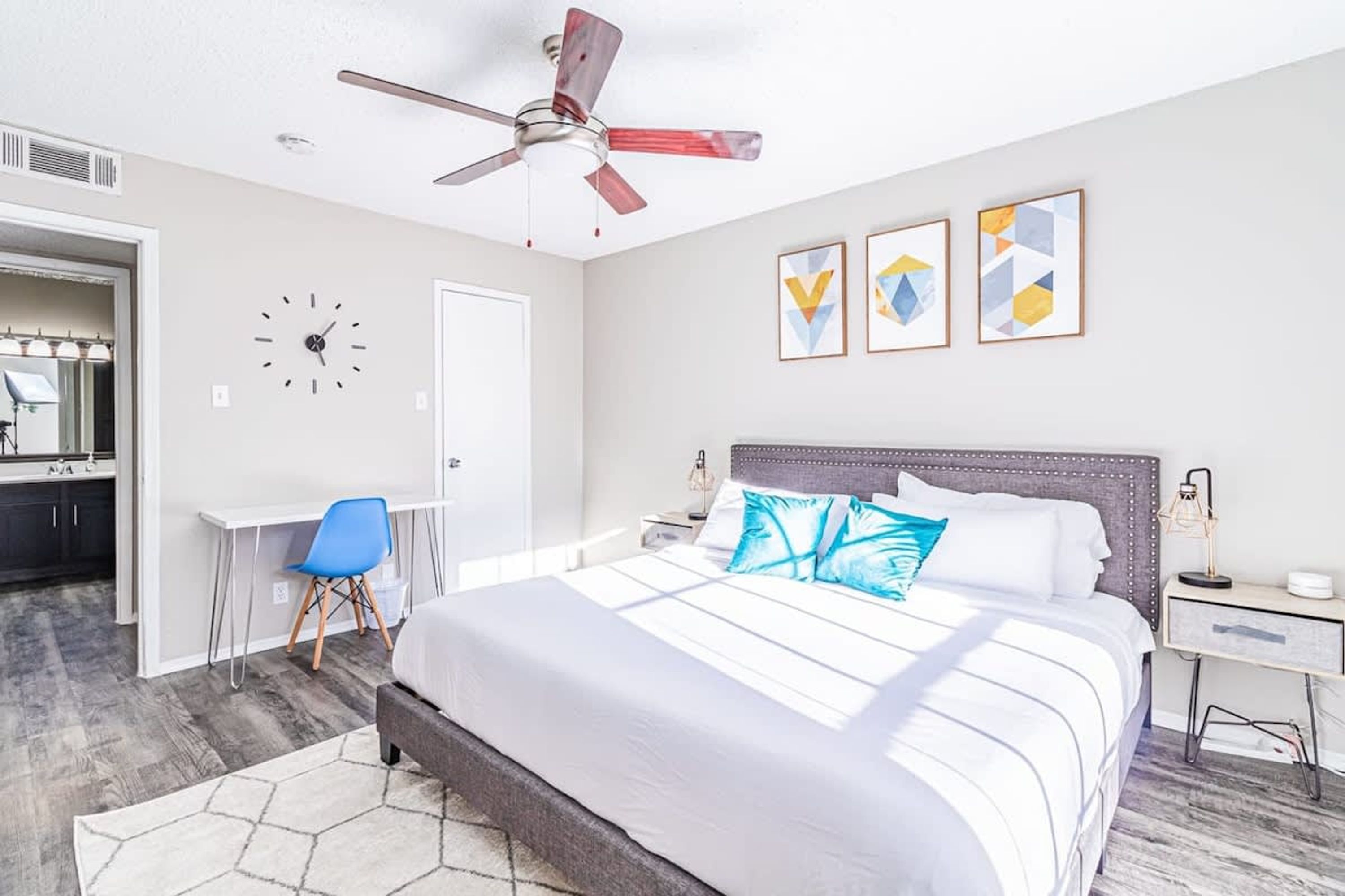 A bedroom with a bed, a desk, a chair, a ceiling fan, and three wall art pieces.