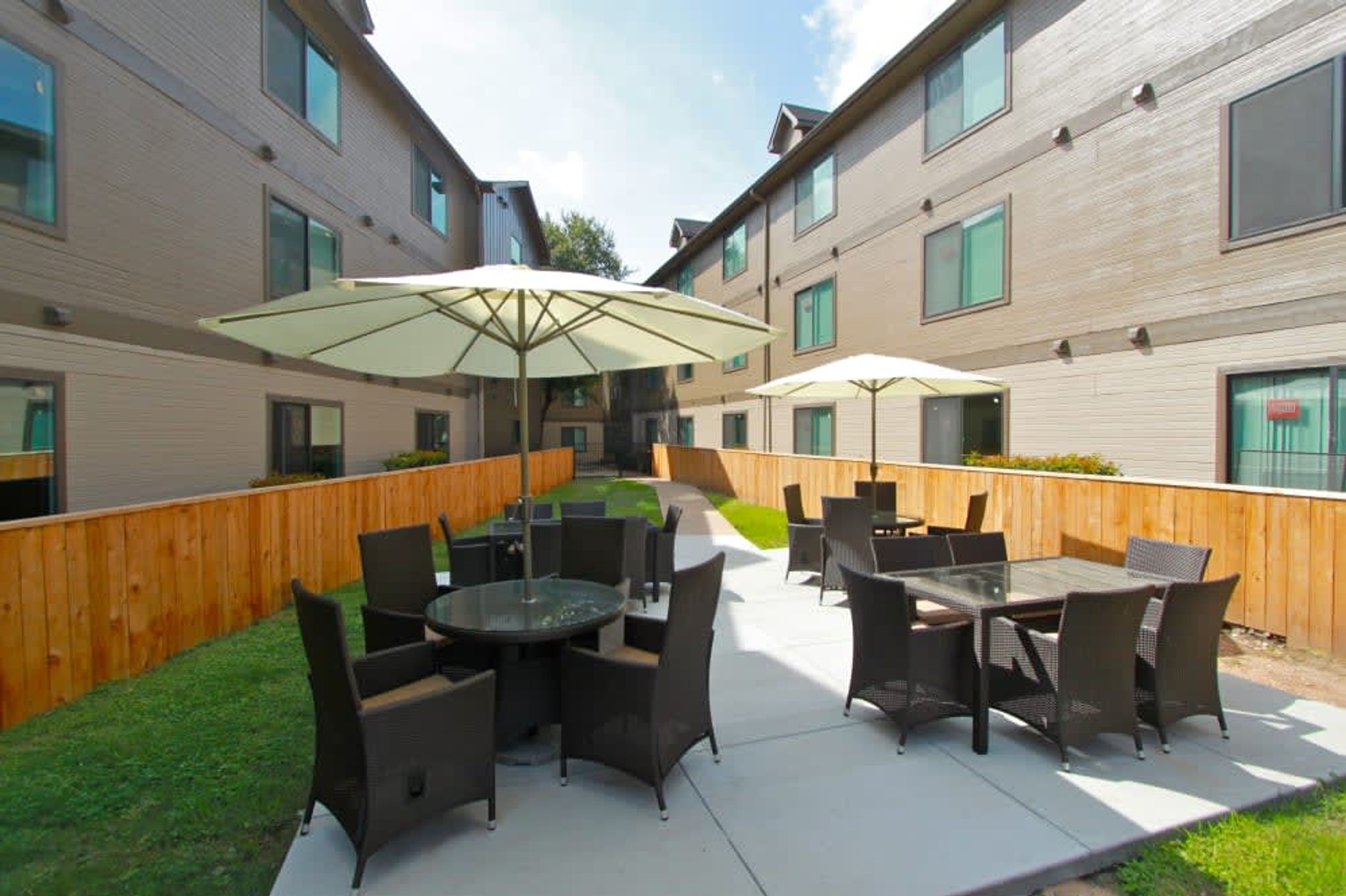 Een terras buiten met tafels en stoelen, omringd door een houten hek en een gebouw met meerdere ramen.