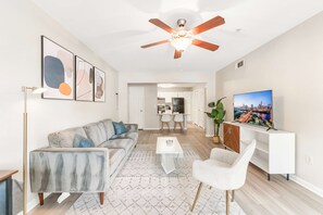 Un salon moderne avec un canapé, une table basse et un téléviseur. On y trouve également un ventilateur de plafond, des œuvres d’art accrochées au mur et, au fond, une cuisine.