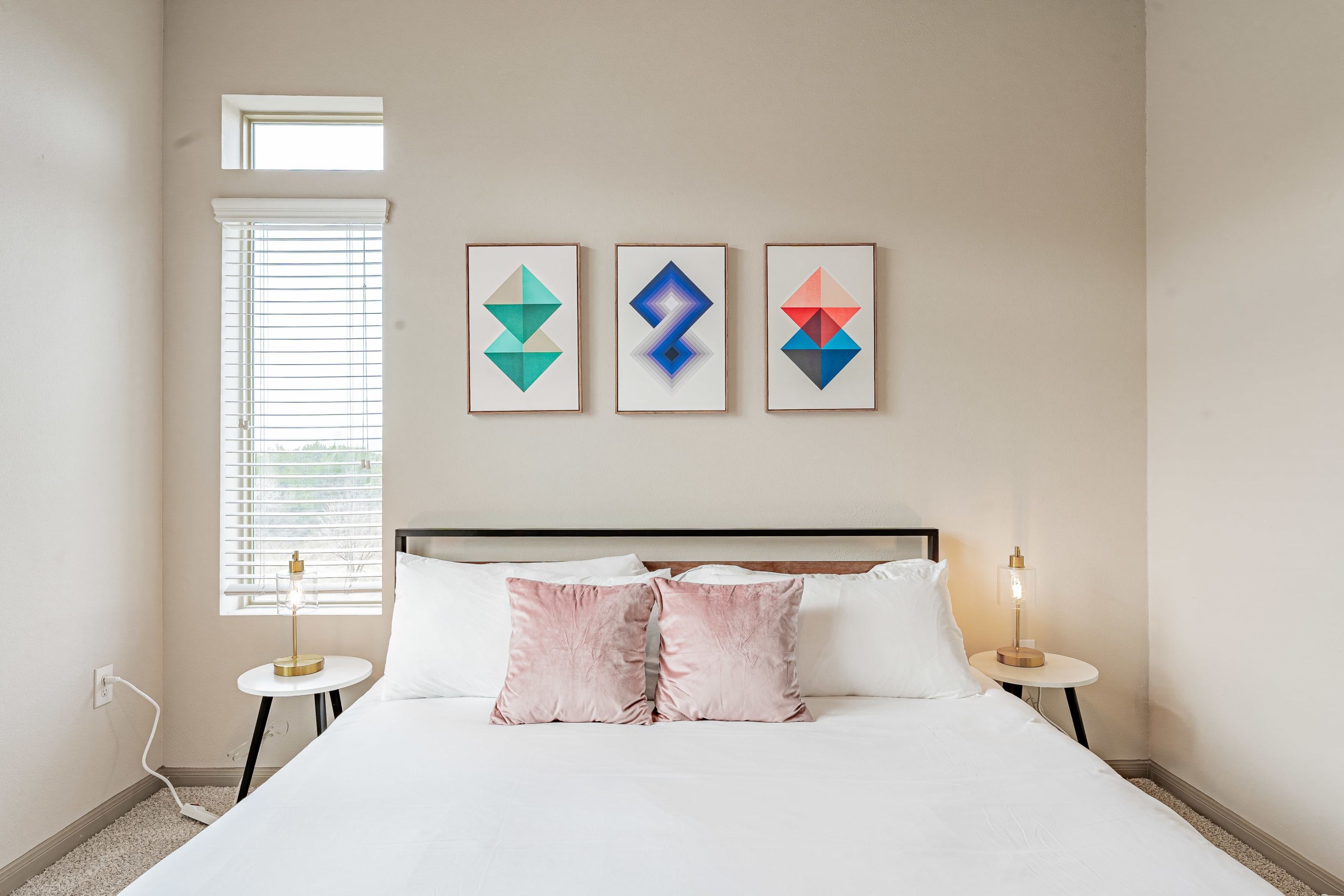 Hotellihuone, jossa on sänky, kaksi yöpöytää, pieni sivupöytä, ikkuna, jossa on sälekaihtimet, ja seinällä kolme kehystettyä, geometrisesti muotoiltua taideteosta.
