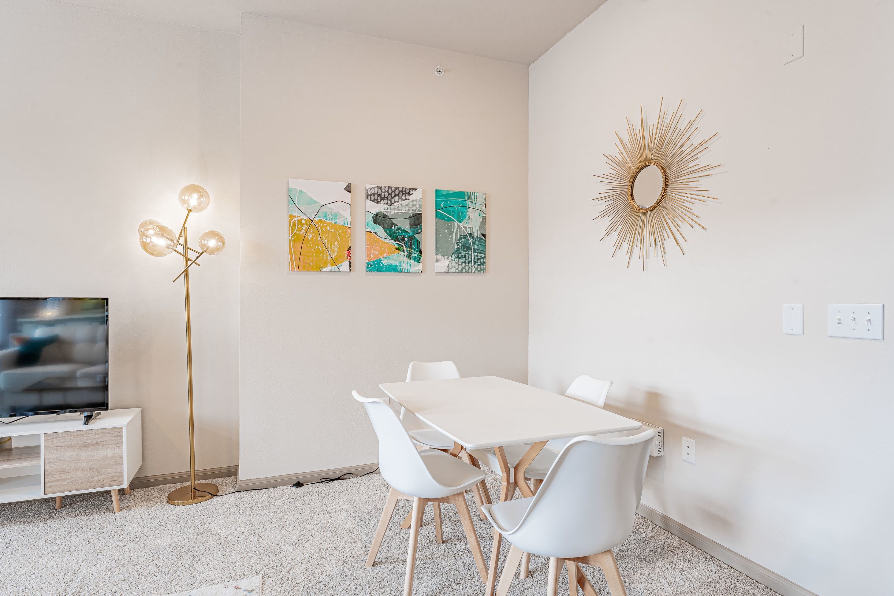 A modern living room with a white table, white chairs, a floor lamp, and abstract wall art.