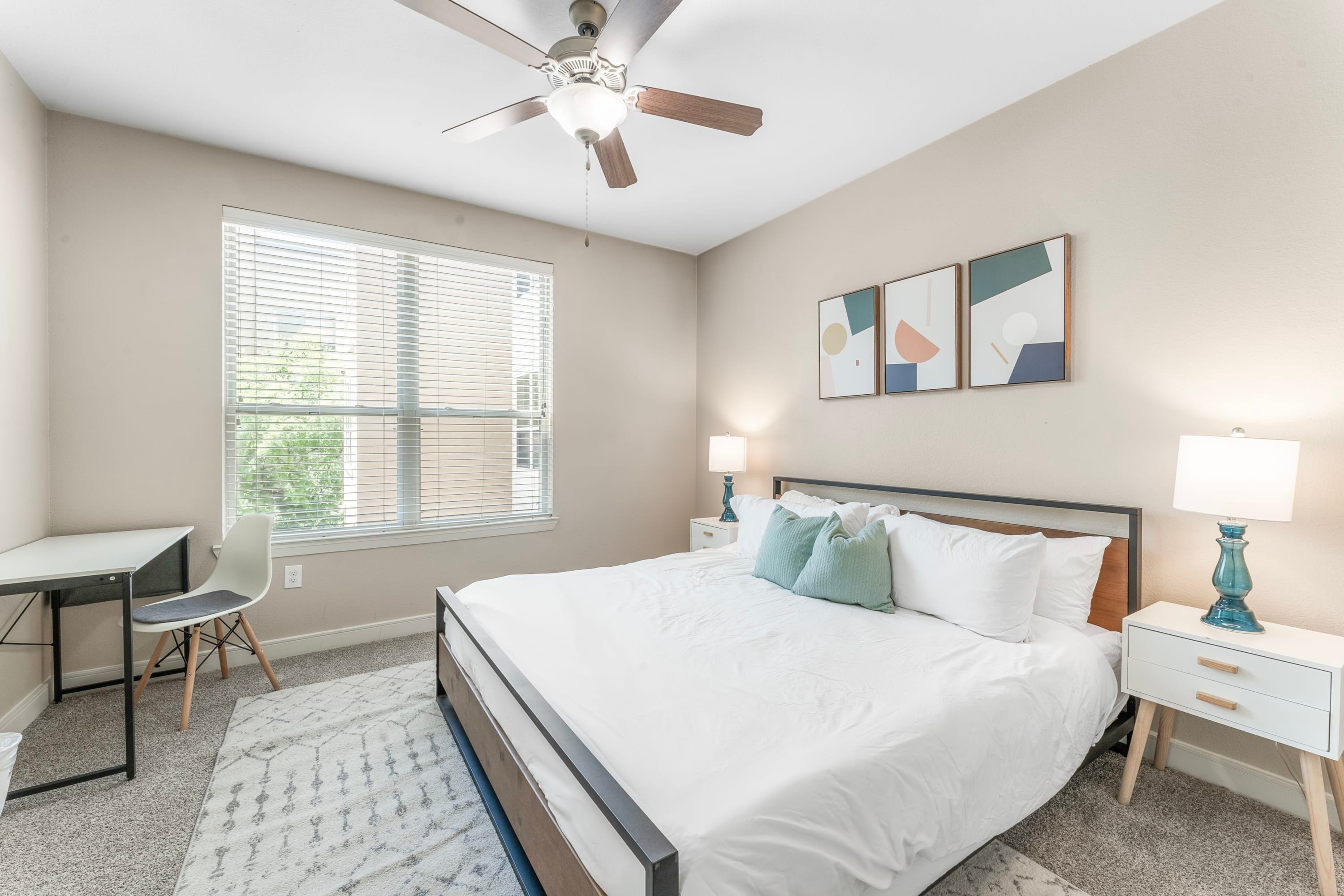 A bedroom with a bed, a desk, a chair, a ceiling fan, and artwork on the wall.