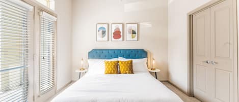 Una habitación de hotel con una cama grande, cabecera azul, ropa de cama blanca y vistas de la ciudad a través de ventanales de gran altura.