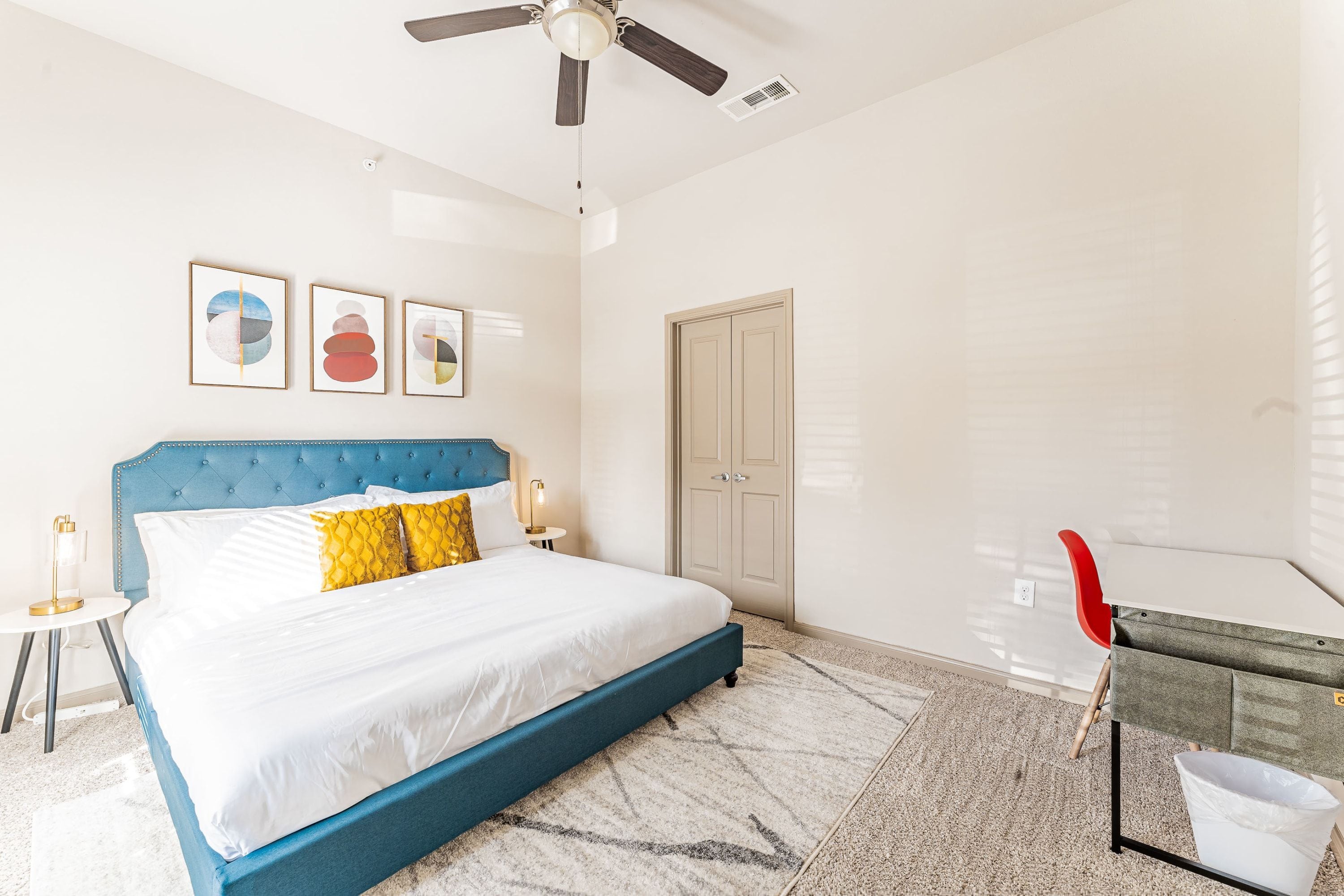 A hotel room with a bed, a ceiling fan, a door, and a small table with a lamp.
