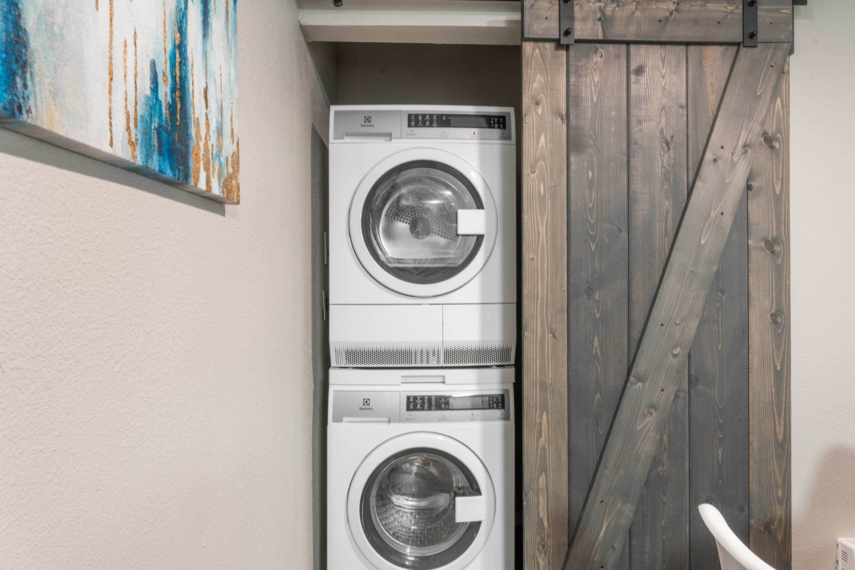 A laundry room with two stacked front-loading washing machines, a wooden door, and a painting on the wall.