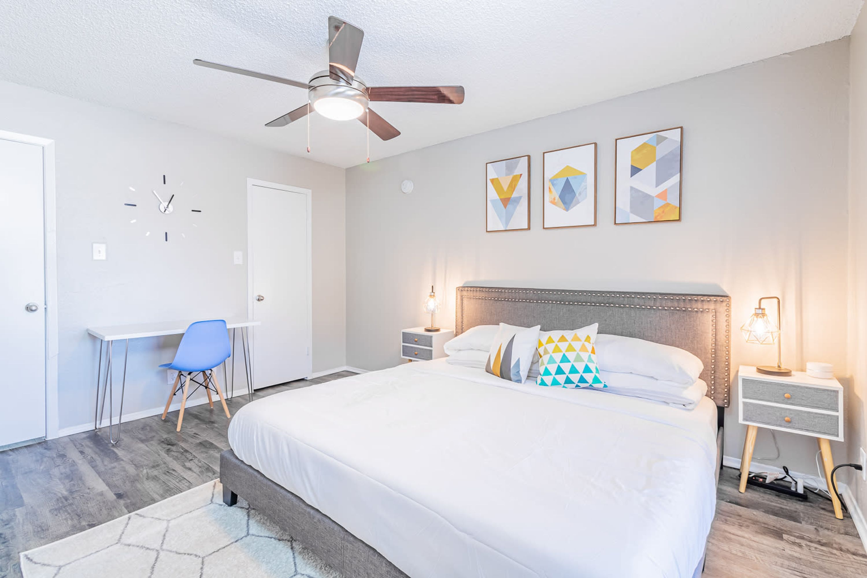 Un dormitorio con cama, escritorio, silla, ventilador de techo y tres obras de arte en la pared.
