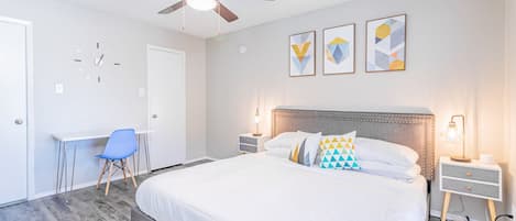 Un dormitorio con cama, escritorio, silla, ventilador de techo y tres obras de arte en la pared.