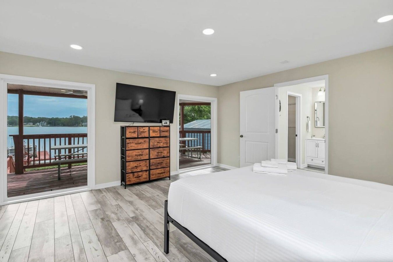Une chambre avec un grand lit, un téléviseur fixé au mur et une commode en bois. La chambre comprend un balcon avec vue sur une étendue d’eau.