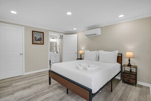 A bedroom with a bed, bedside tables, an air conditioning unit, and a bathroom visible through an open door.