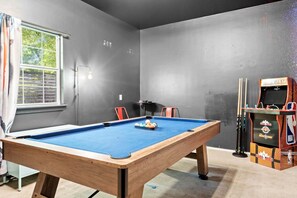 A pool table with balls, a pool cue rack, and a basketball arcade machine in a room with grey walls. - Two Game Rooms, Two Fire Pit Courtyards & Two Kitchens | Room for 20 in East ATX (Austin)