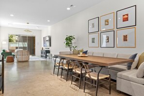 A modern living room with a dining area, featuring a wooden table, chairs, a sofa, and framed artwork on the wall. - Two Game Rooms, Two Fire Pit Courtyards & Two Kitchens | Room for 20 in East ATX (Austin)