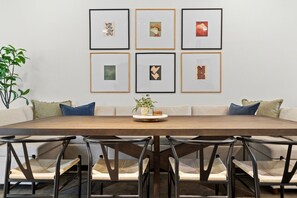 A dining area with a long table, six chairs, and six framed artworks on the wall. - Two Game Rooms, Two Fire Pit Courtyards & Two Kitchens | Room for 20 in East ATX (Austin)