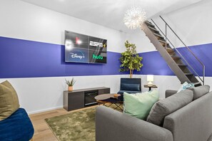 A modern living room with a flat-screen TV mounted on the wall, a staircase, and a potted plant. - Two Game Rooms, Two Fire Pit Courtyards & Two Kitchens | Room for 20 in East ATX (Austin)