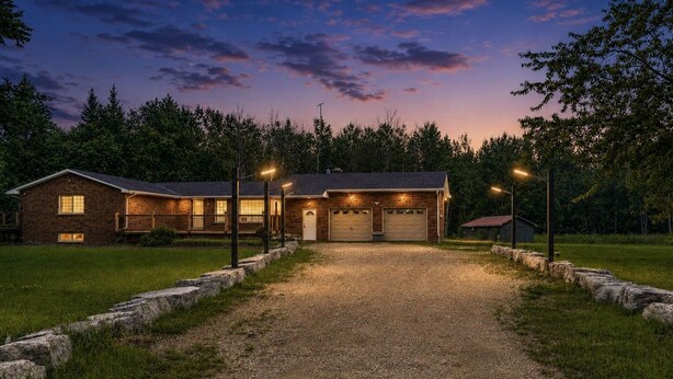 A two-story house with a gravel driveway leading to a garage, surrounded by a stone wall and trees.