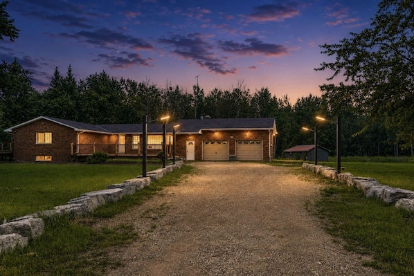 A two-story house with a gravel driveway leading to a garage, surrounded by a stone wall and trees.