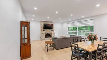 A modern living room with a fireplace, a large TV, a sofa, and a dining area with a wooden table and chairs.