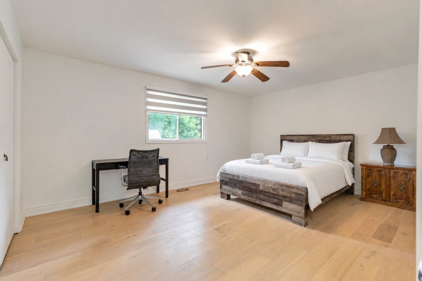 A bedroom with a bed, a desk, a chair, a ceiling fan, and a window.
