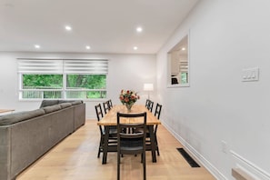A dining area with a wooden table, chairs, a sofa, and a window with blinds.