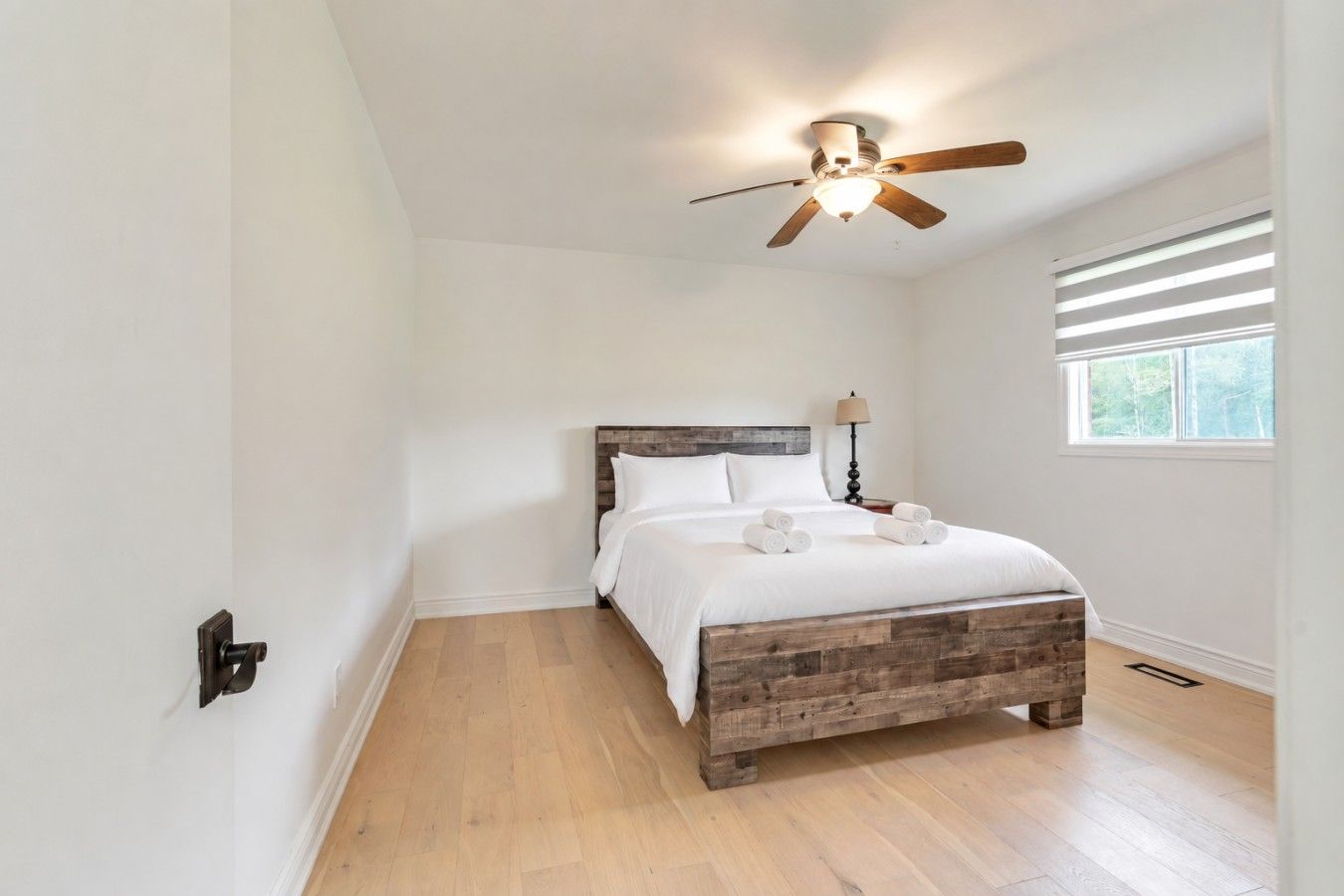 Un dormitorio con cama de madera, ventilador de techo, ventana con persianas y mesita de noche con lámpara.