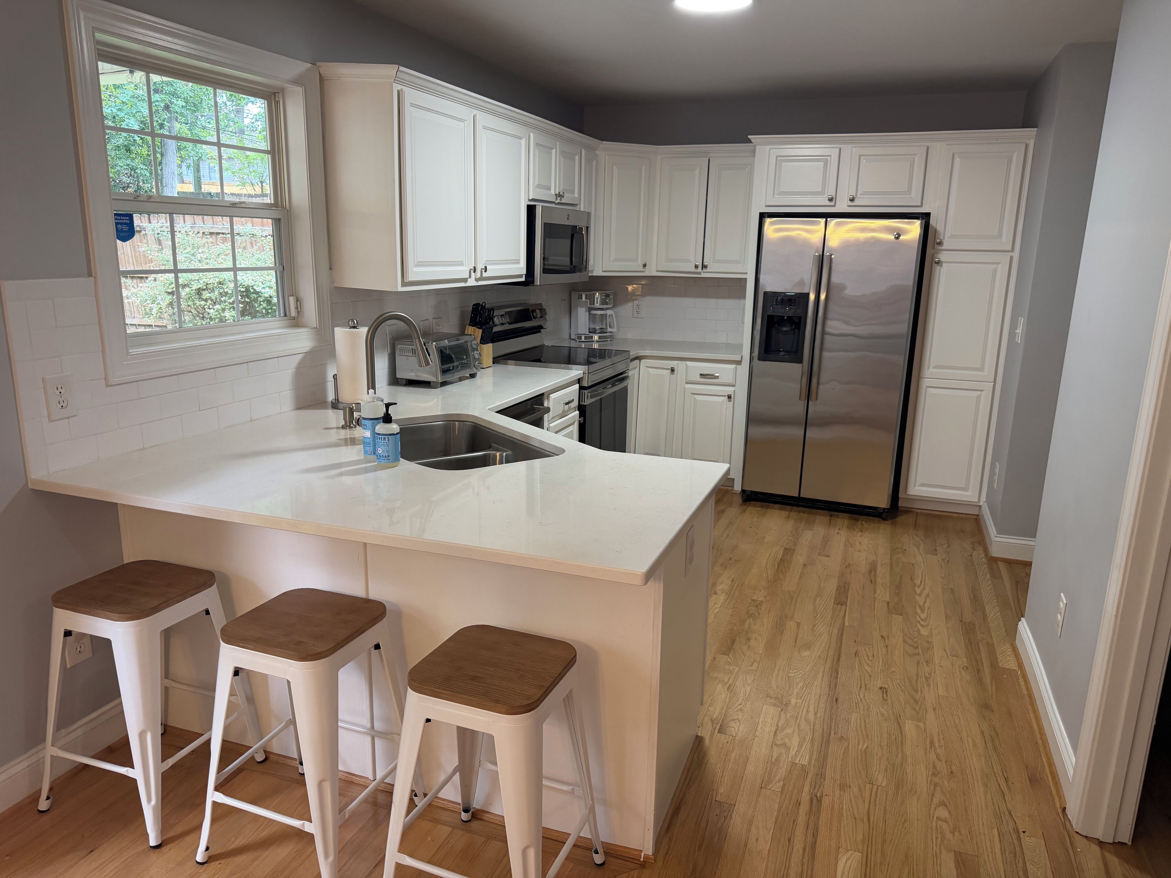 A kitchen with white cabinets, a stainless steel refrigerator, a double sink, and wooden stools.