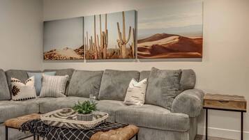 A living room with a grey sofa, patterned cushions, a small side table, and a wall-mounted painting of cacti.