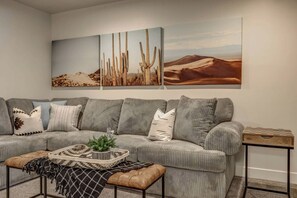 Un salon avec un canapé gris, des coussins à motifs, une petite table d’appoint et un tableau représentant des cactus accroché au mur.